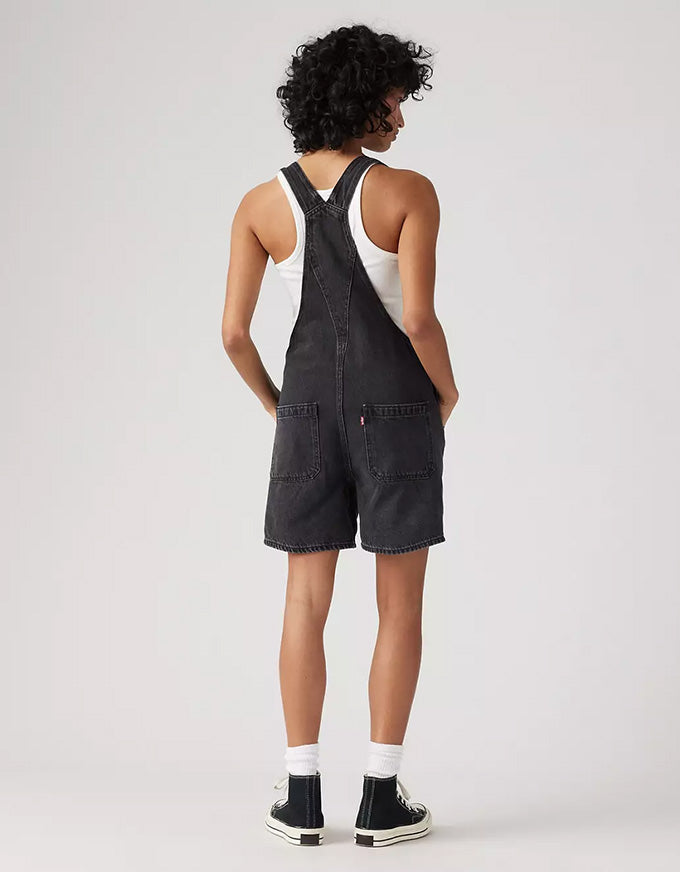 A model shown from the back wearing dark gray denim overall shorts over a white tank top, paired with white socks and black high-top sneakers, against a light gray studio background.