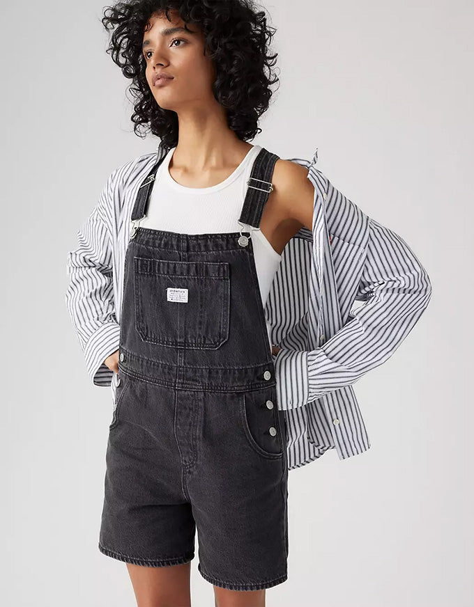 A model wearing dark gray denim overall shorts with a white tank top, a striped button-up shirt, and a black hairpiece, photographed against a light gray studio background.
