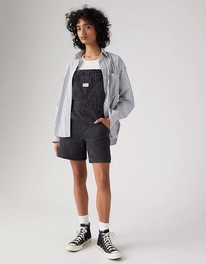A model wearing a striped button-down shirt over a black denim overall outfit, styled with a white t-shirt, white socks, and black high-top sneakers against a light gray studio background.