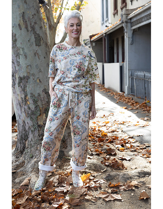 A woman stands outdoors on a leaf-strewn sidewalk beside a large tree, wearing a matching two-piece pajama set with a light beige base and a multicolor floral paisley print. The set includes a short-sleeve top and drawstring pants, paired with white cuffed boots.