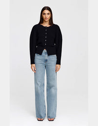 A woman stands against a light gray studio background wearing a black long-sleeve button-front top and light-wash wide-leg jeans.