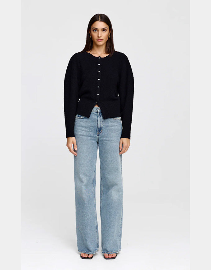 A woman stands against a light gray studio background wearing a black long-sleeve button-front top and light-wash wide-leg jeans.