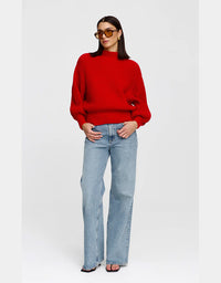 A woman modeling a bright red ribbed knit mock-neck sweater with light-wash wide-leg jeans and sunglasses, standing against a light gray studio background.