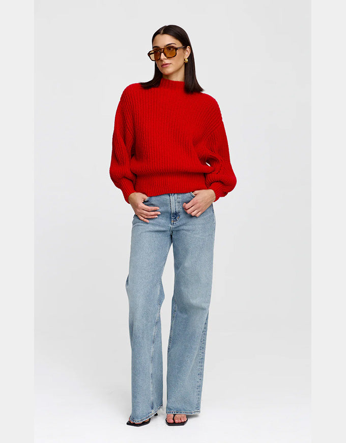 A woman modeling a bright red ribbed knit mock-neck sweater with light-wash wide-leg jeans and sunglasses, standing against a light gray studio background.