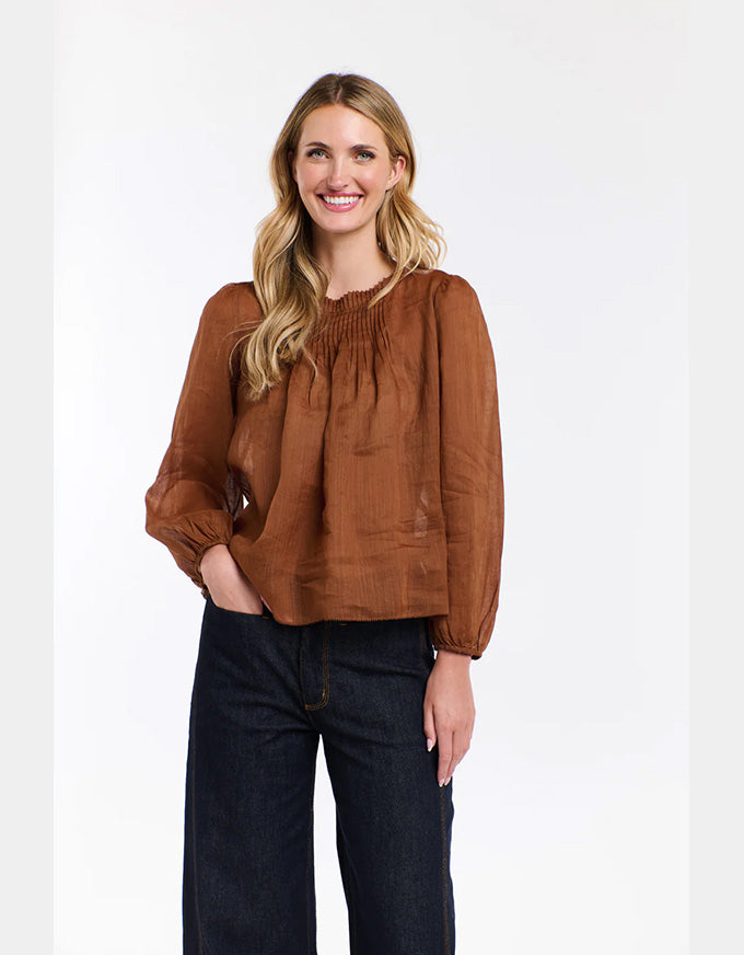 A woman modeling a brown long-sleeve linen blouse with a smocked neckline and dark wide-leg jeans, standing against a plain white studio background.