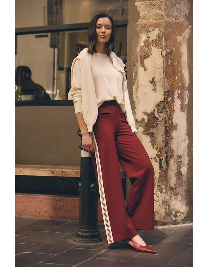 Woman in a white sweater and red pants standing against a textured wall.