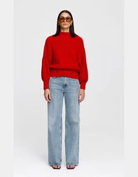 A woman modeling a bright red ribbed knit mock-neck sweater with balloon sleeves, paired with light-wash wide-leg jeans and sunglasses, against a plain light gray background.