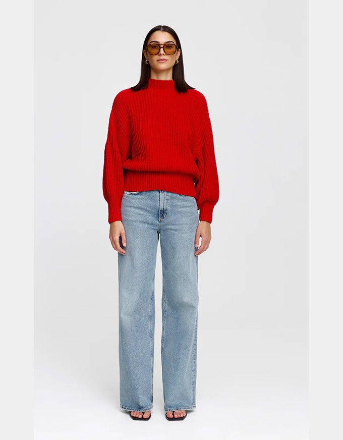 A woman modeling a bright red ribbed knit mock-neck sweater with balloon sleeves, paired with light-wash wide-leg jeans and sunglasses, against a plain light gray background.