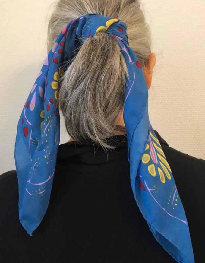 Person wearing a blue scarf with colorful patterns around their head against a plain background