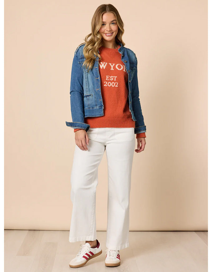 Woman wearing a red sweater with 'NYC' text, blue denim jacket, white pants, and white sneakers on a beige background