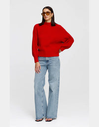 A woman modeling a bright red ribbed knit sweater with balloon sleeves, paired with light-wash wide-leg jeans and sunglasses, against a plain light gray studio background.