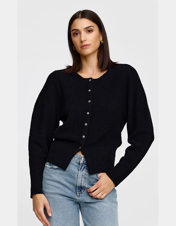 A woman wearing a black long-sleeve knit cardigan with a button-down front, paired with light-wash blue jeans, against a plain light gray studio background.