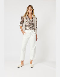 A woman modeling a stylish outfit consisting of a leopard print blouse, a light beige vest, white cropped trousers, and cream-colored loafers, set against a plain light gray studio background.