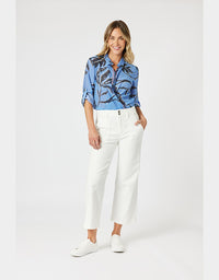 A woman modeling a light blue button-up shirt with a dark abstract print, paired with white cropped trousers and white sneakers, against a light gray studio background.