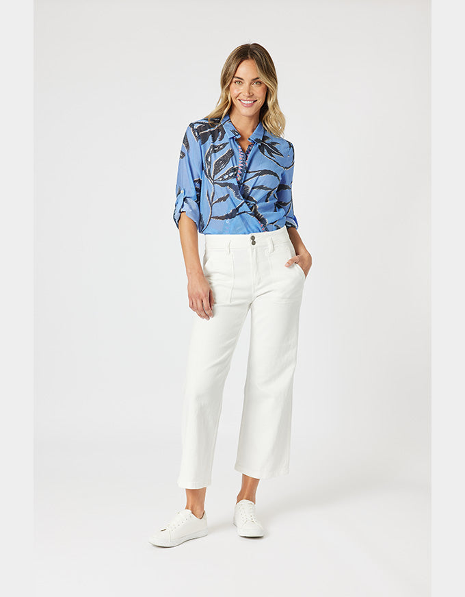 A woman modeling a light blue button-up shirt with a dark abstract print, paired with white cropped trousers and white sneakers, against a light gray studio background.