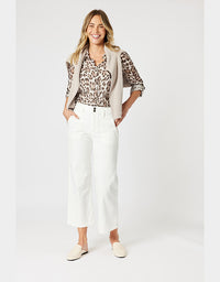A woman modeling a beige sleeveless vest over a leopard-print blouse, paired with white cropped pants and cream loafers, against a light gray studio background.