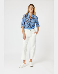 A woman modeling a blue short-sleeve button-up shirt with a black abstract print, paired with white cropped pants and white sneakers, against a light gray studio background.