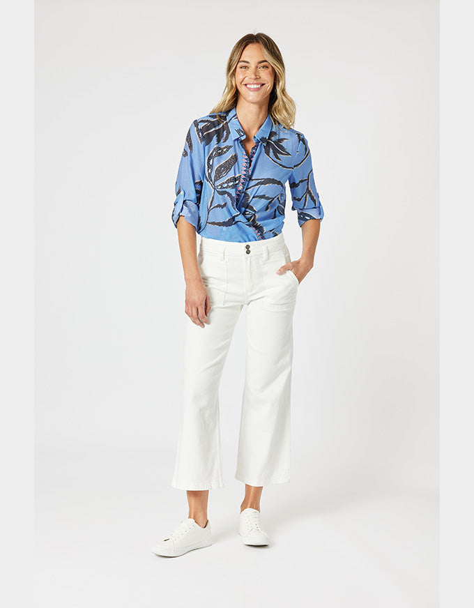 A woman modeling a blue short-sleeve button-up shirt with a black abstract print, paired with white cropped pants and white sneakers, against a light gray studio background.