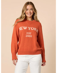 Woman wearing an orange sweater with 'New York Est 2002' text on a beige background