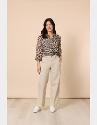 A woman stands in a studio wearing a leopard-print blouse with three-quarter sleeves, beige wide-leg trousers, and white sneakers against a warm beige backdrop.