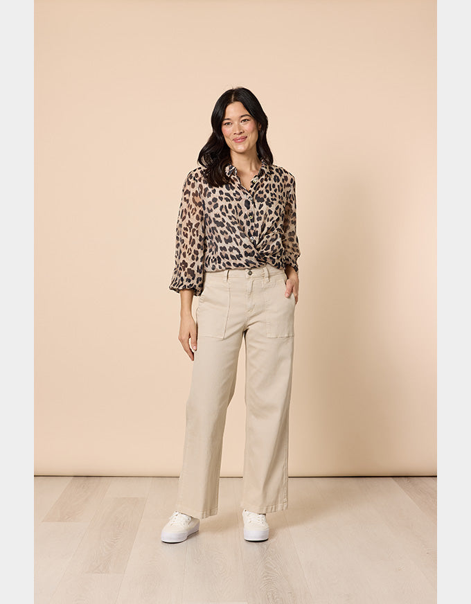 A woman stands in a studio wearing a leopard-print blouse with three-quarter sleeves, beige wide-leg trousers, and white sneakers against a warm beige backdrop.