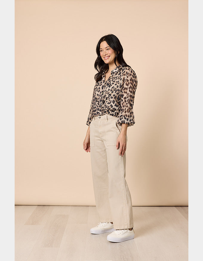 A woman modeling a leopard-print long-sleeve blouse and beige wide-leg trousers with white sneakers, standing against a solid beige studio backdrop.