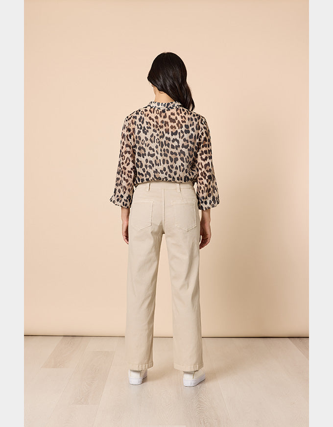 A woman is shown from behind wearing a leopard-print long-sleeve blouse and beige wide-leg trousers, standing against a solid beige studio background.