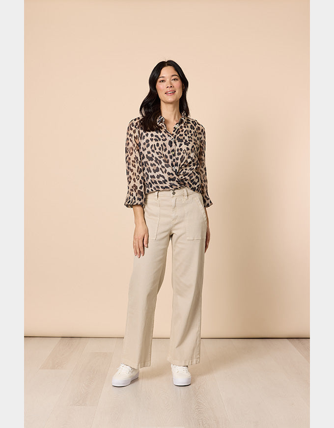 A woman stands in a studio wearing a leopard-print button-up blouse with three-quarter sleeves, beige wide-leg trousers, and white sneakers against a warm beige backdrop.