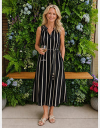 A woman stands outdoors in front of a lush green garden wall, wearing a sleeveless black midi dress with vertical cream stripes and a matching tie belt. She is holding a glass of white wine and wearing beige slide sandals.