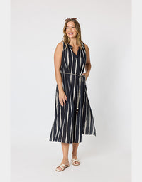 A woman modeling a sleeveless midi dress with vertical black and white stripes, a tie neckline, and a matching striped belt with a chain detail, set against a plain light gray studio background.