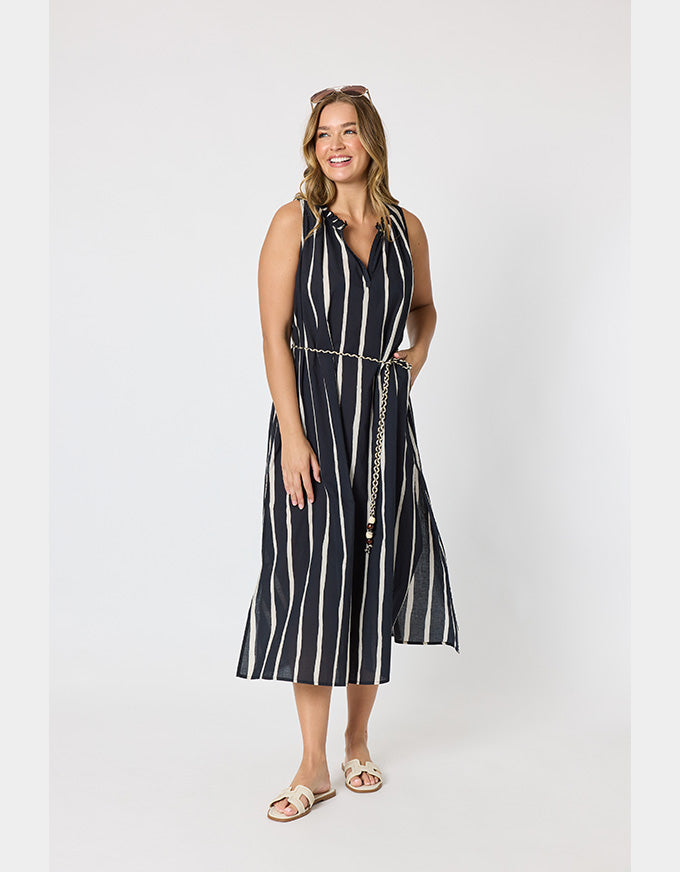 A woman modeling a sleeveless midi dress with vertical black and white stripes, a tie neckline, and a matching striped belt with a chain detail, set against a plain light gray studio background.