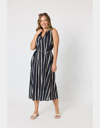 A woman modeling a sleeveless midi dress with vertical black and white stripes, featuring a V-neckline, a matching thin belt with a gold-tone chain detail, and light-colored flat sandals, set against a plain light gray studio background.