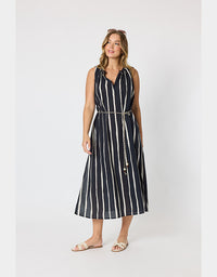 A woman modeling a sleeveless midi dress with vertical white stripes on a black background, featuring a V-neckline, a matching tie belt with a chain detail, and flat slide sandals, set against a plain light gray studio background.
