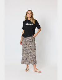 A woman modeling a black short-sleeve t-shirt with 'Amour' written in white script across the chest, paired with a leopard-print midi skirt and beige slide sandals, against a light gray studio background.