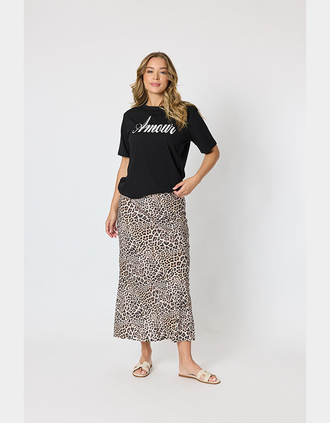 A woman modeling a black short-sleeve t-shirt with 'Amour' written in white script across the chest, paired with a leopard-print midi skirt and beige slide sandals, against a light gray studio background.