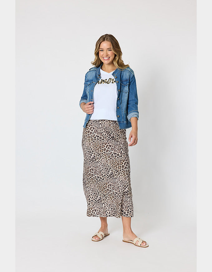 A woman modeling a leopard print midi skirt, styled with a white t-shirt, a blue denim jacket, and beige slide sandals, against a plain light gray studio background.