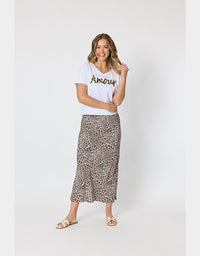 A woman modeling a white graphic t-shirt and a brown leopard-print midi skirt against a plain light gray studio background.