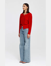 A woman modeling a bright red long-sleeve knit cardigan with a buttoned front, paired with light-wash wide-leg jeans and flat sandals, against a plain light gray studio background.