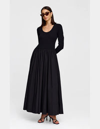 A woman modeling a long-sleeve black maxi dress with a scoop neckline and a gathered waist, paired with black strappy heels and sunglasses, against a light gray studio background.