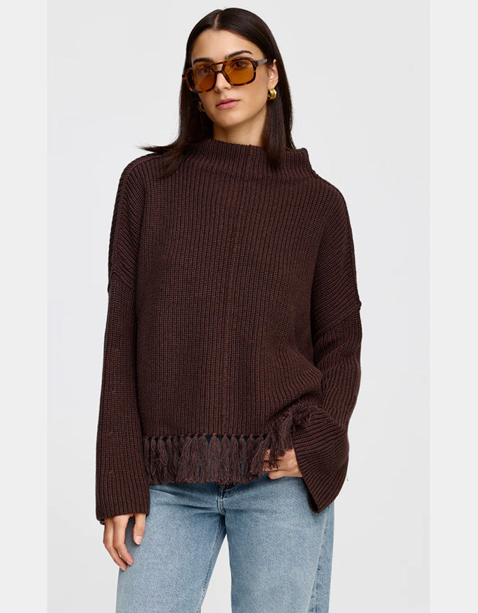 A woman wearing a dark brown ribbed knit sweater with fringe trim and sunglasses, paired with light blue jeans, against a light gray background.