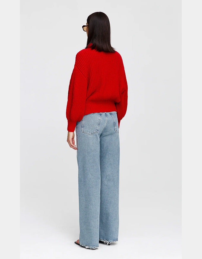 A woman is shown from behind wearing a bright red knit sweater and light blue wide-leg jeans against a plain light gray studio background.