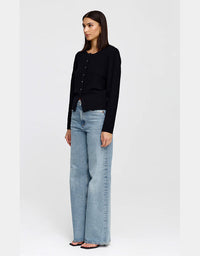 A woman modeling a black long-sleeve button-front cardigan with light-wash wide-leg jeans, standing against a plain light gray studio background.