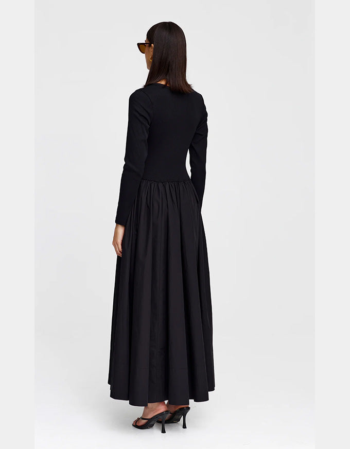A woman wearing a long black dress with a fitted long-sleeve bodice and a full, flowing skirt, shown from the back against a plain light gray studio background.