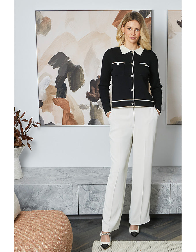 Woman wearing a black jacket with white buttons and white pants in a room with abstract art on the wall.