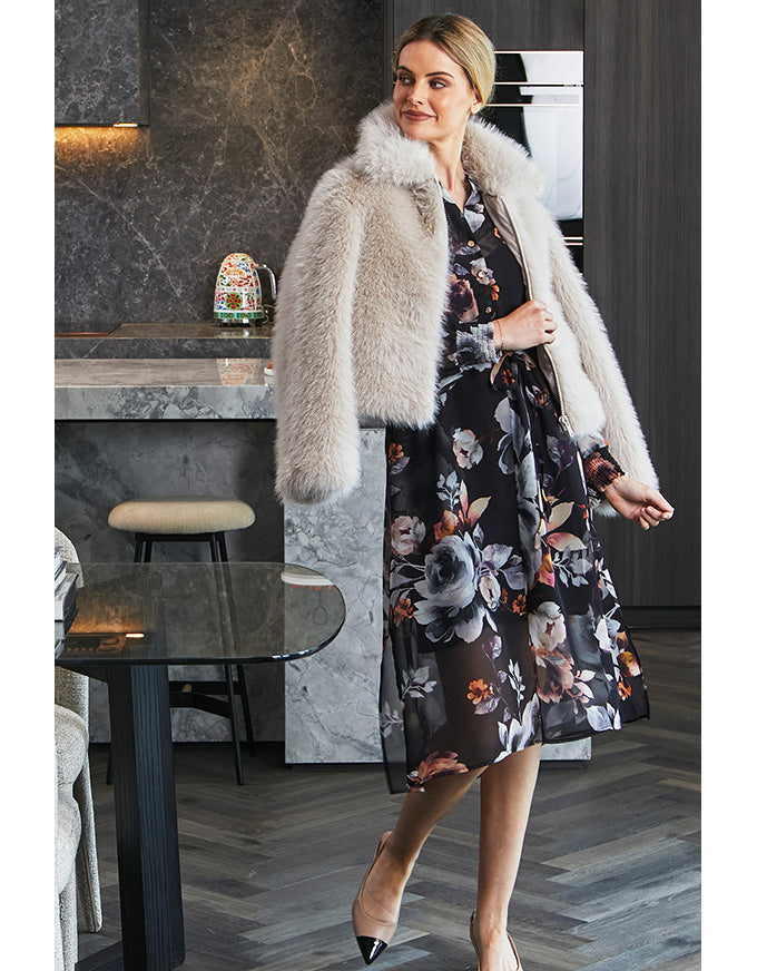 Woman in a floral dress and fur coat standing in a modern kitchen.