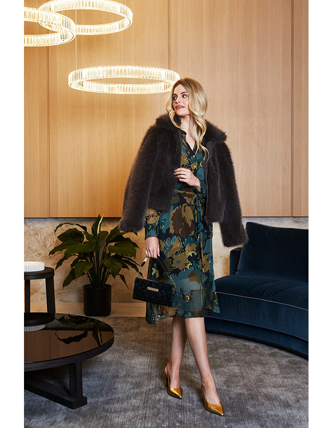 Woman in a stylish outfit standing in a modern interior setting with wooden walls and a blue sofa.