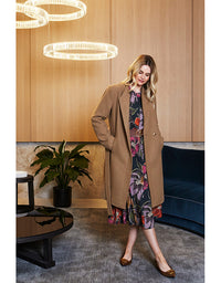 Woman in a beige coat and floral dress standing in a modern interior setting.