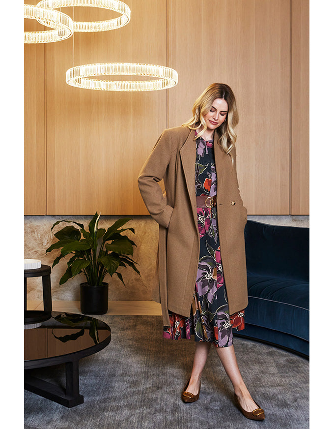 Woman in a beige coat and floral dress standing in a modern interior setting.