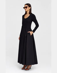 A woman modeling a long-sleeve black maxi dress with a scoop neckline and side pockets, paired with black strappy heels and sunglasses, against a light gray studio background.