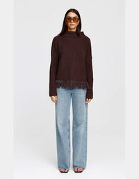 A model wearing a dark brown ribbed knit sweater with fringe trim, paired with light-wash wide-leg jeans and sunglasses, standing against a light gray studio background.